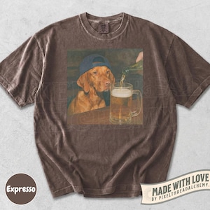 K&ouml;nnte beinhalten: Ein braunes Kurzarm-T-Shirt mit einem Grafikdruck eines braunen Hundes, der eine blaue Baseballkappe tr&auml;gt und neben einem mit Bier gef&uuml;llten Glaskrug sitzt. Das T-Shirt hat einen Rundhalsausschnitt und ein verwaschenes braunes Stofffinish.