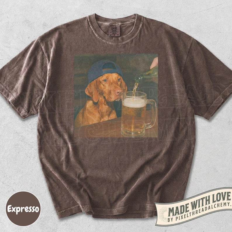 K&ouml;nnte beinhalten: Ein braunes Kurzarm-T-Shirt mit einem Grafikdruck eines braunen Hundes, der eine blaue Baseballkappe tr&auml;gt und neben einem mit Bier gef&uuml;llten Glaskrug sitzt. Das T-Shirt hat einen Rundhalsausschnitt und ein verwaschenes braunes Stofffinish.