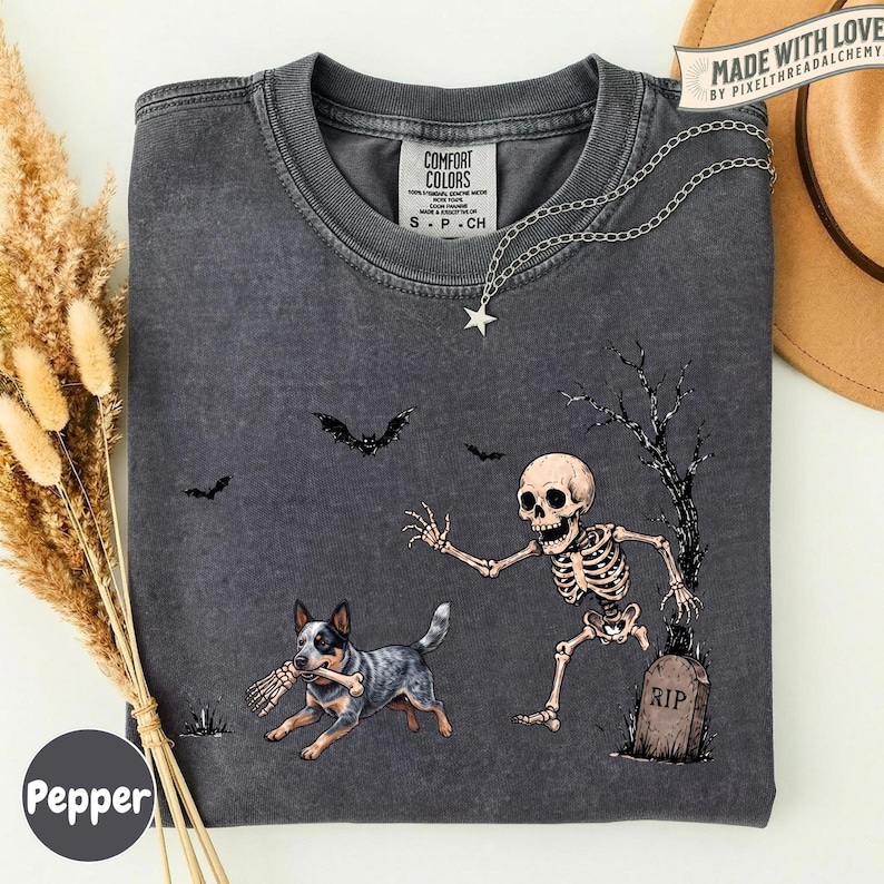 May include: A charcoal gray t-shirt featuring a graphic of a skeleton running from a tombstone while a dog carries a bone in its mouth. Bats fly in the background near a bare tree. The shirt has a crew neck and a Comfort Colors brand label.