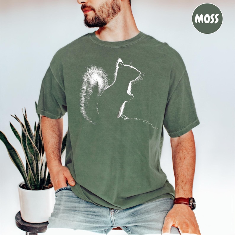 A man wearing a green t-shirt with a white silhouette of a squirrel is sitting in front of a potted plant.