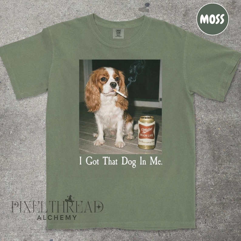 A t-shirt with a graphic design featuring a brown and white dog sitting on a wooden surface, holding a can of Miller High Life beer.