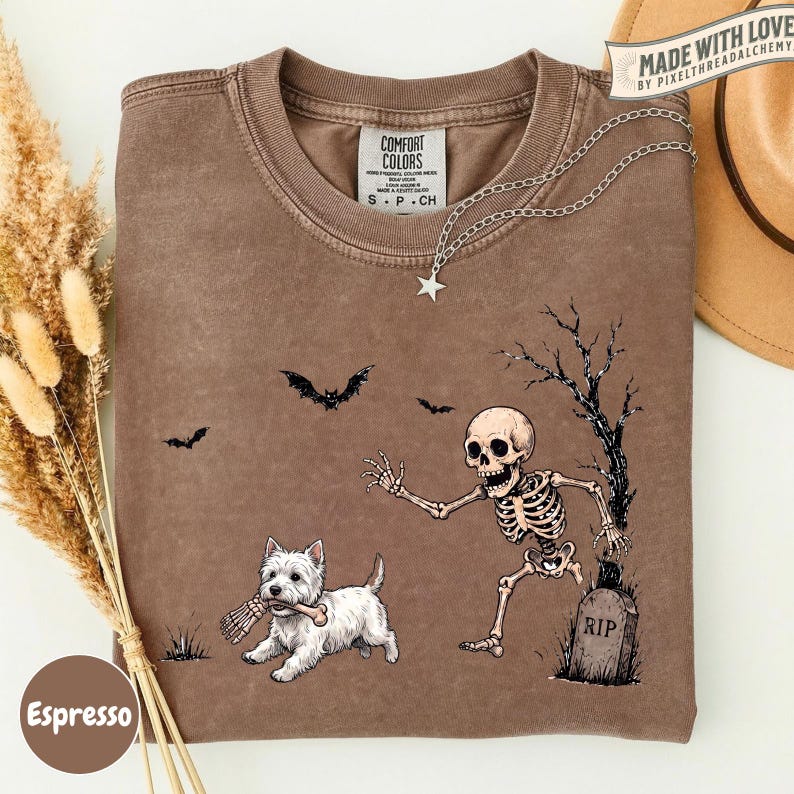 May include: A brown crewneck t-shirt featuring a graphic of a white West Highland Terrier dog holding a bone, a skeleton reaching toward the dog, a tombstone labeled RIP, and flying bats. The shirt has a Comfort Colors brand neck label.