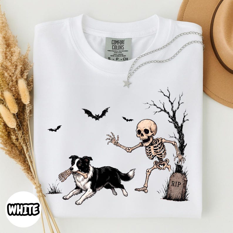May include: A white t-shirt featuring a graphic of a black and white dog holding a bone in its mouth, running away from a human skeleton near a tombstone marked RIP. The design includes a bare tree and three flying bats. The shirt has a Comfort Colors label.