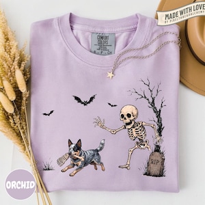 May include: A light purple t-shirt featuring a graphic print of a skeleton running from a tombstone marked RIP, while a blue heeler dog runs with a bone in its mouth. Bats fly in the background near a bare tree. The shirt has a Comfort Colors label.