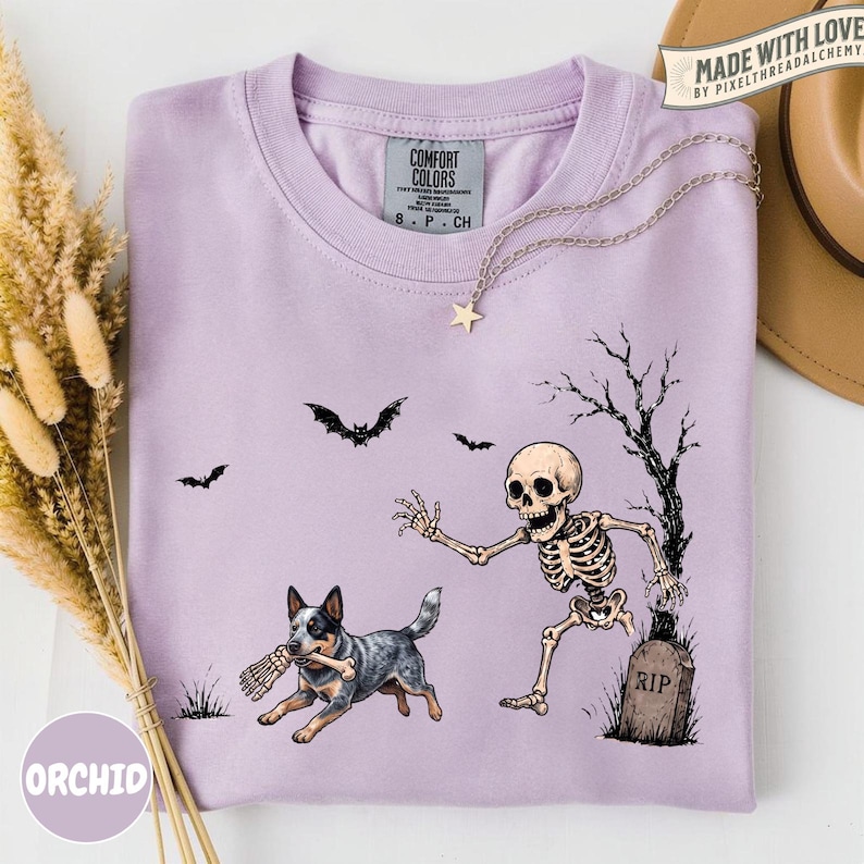 May include: A light purple t-shirt featuring a graphic print of a skeleton running from a tombstone marked RIP, while a blue heeler dog runs with a bone in its mouth. Bats fly in the background near a bare tree. The shirt has a Comfort Colors label.