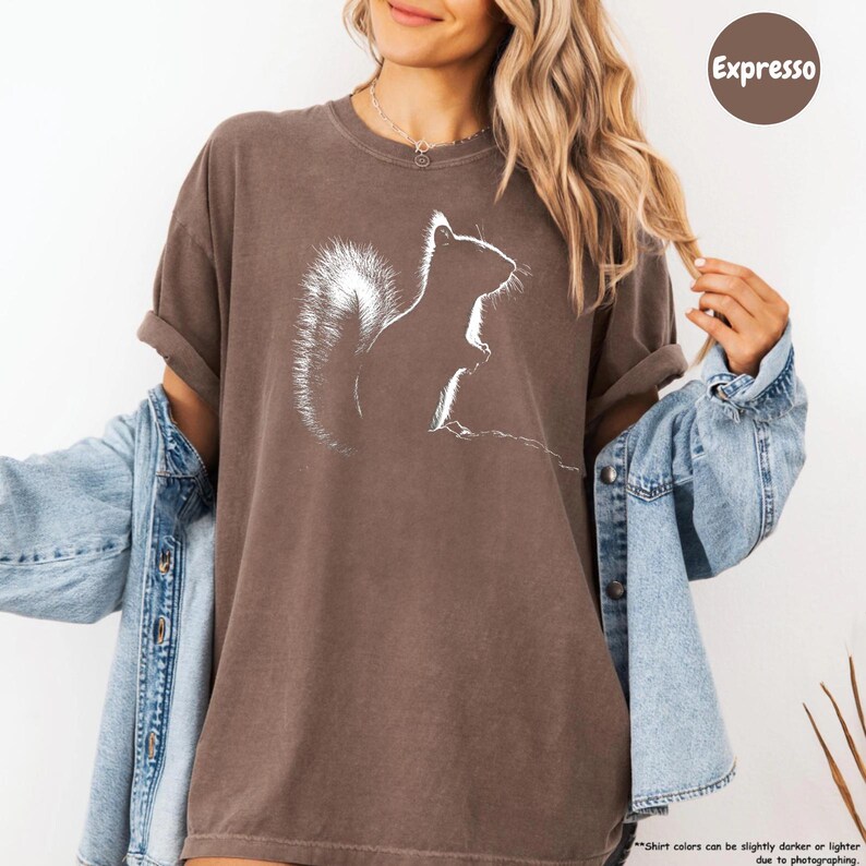 A woman wearing a brown t-shirt with a white squirrel graphic and a denim jacket, smiling and posing for the camera.