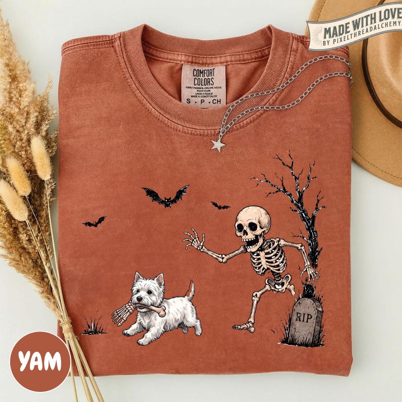 May include: A rust-colored t-shirt featuring a graphic of a skeleton reaching toward a white West Highland Terrier holding a bone in its mouth. The design includes a tombstone labeled RIP, a bare tree, and two bats. A silver star necklace rests on the collar.
