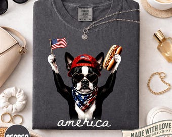 May include: A charcoal grey t-shirt featuring a graphic of a Boston Terrier wearing a red baseball cap, heart-shaped sunglasses, and an American flag bandana. The dog holds a small American flag and a hot dog. The word america is printed below.