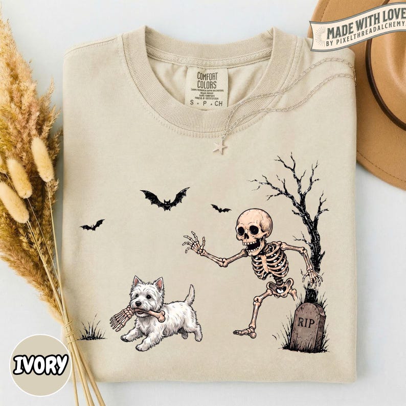 May include: A beige crewneck t-shirt featuring a graphic print of a white West Highland Terrier dog holding a bone in its mouth, running away from a skeleton emerging from a grave near a bare tree, with three black bats flying above.