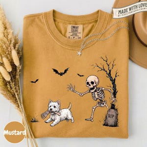 May include: A mustard yellow t-shirt featuring a graphic print of a white West Highland Terrier dog holding a bone in its mouth, a skeleton reaching toward the dog, a tombstone marked RIP, a bare tree, and two flying bats against a plain background.