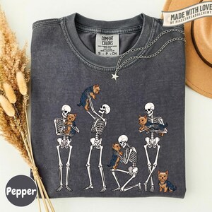 May include: A charcoal gray t-shirt featuring a graphic of four white skeletons interacting with small brown and black Yorkshire Terrier dogs. The shirt has a crew neckline and a Comfort Colors brand label. The design shows skeletons holding, petting, and standing near the dogs.