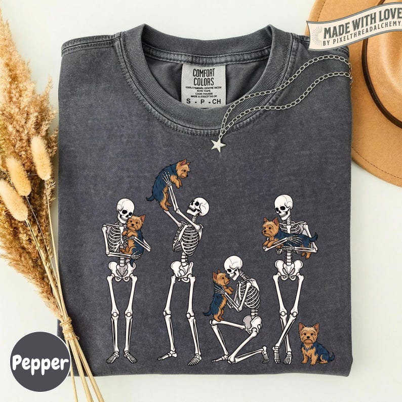 May include: A charcoal gray t-shirt featuring a graphic of four white skeletons interacting with small brown and black Yorkshire Terrier dogs. The shirt has a crew neckline and a Comfort Colors brand label. The design shows skeletons holding, petting, and standing near the dogs.
