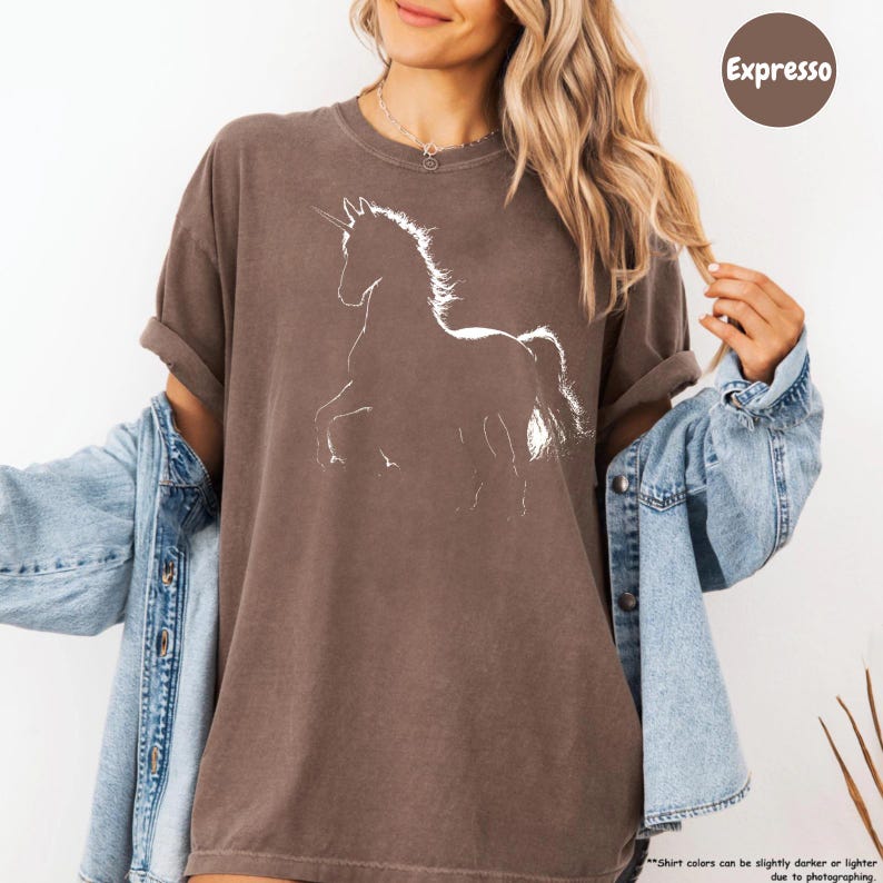 A woman wearing a brown t-shirt with a white unicorn graphic and a denim jacket, smiling and posing for the camera.