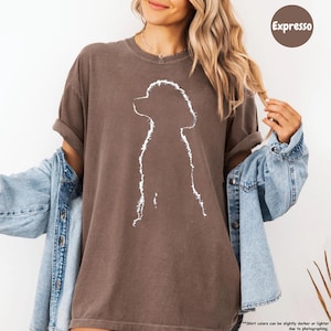 A woman wearing a brown t-shirt with a white silhouette of a dog, paired with a denim jacket, posing against a white background.
