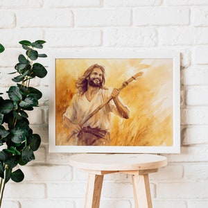 May include: A framed watercolor print of a smiling person holding a staff, set against a golden wheat field. The artwork is in a white frame, displayed on a wooden stool against a white brick wall. Green leafy branches are to the left.