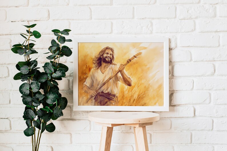 May include: A framed watercolor print of a smiling person holding a staff, set against a golden wheat field. The artwork is in a white frame, displayed on a wooden stool against a white brick wall. Green leafy branches are to the left.