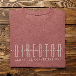 May include: A folded, dusty rose-colored t-shirt with the word "DIRECTOR" in a vertical, sans-serif font. Below it, the words "foundations essentials" and "CLASSICAL CONVERSATIONS" are printed in a script and sans-serif font, respectively. The shirt rests on a wooden surface.