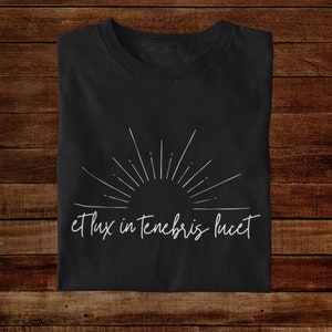 K&ouml;nnte beinhalten: Schwarzes T-Shirt mit einem wei&szlig;en Sonnenstrahlen-Design und dem Text "et lux in tenebris lucet" in handschriftlicher Schrift.