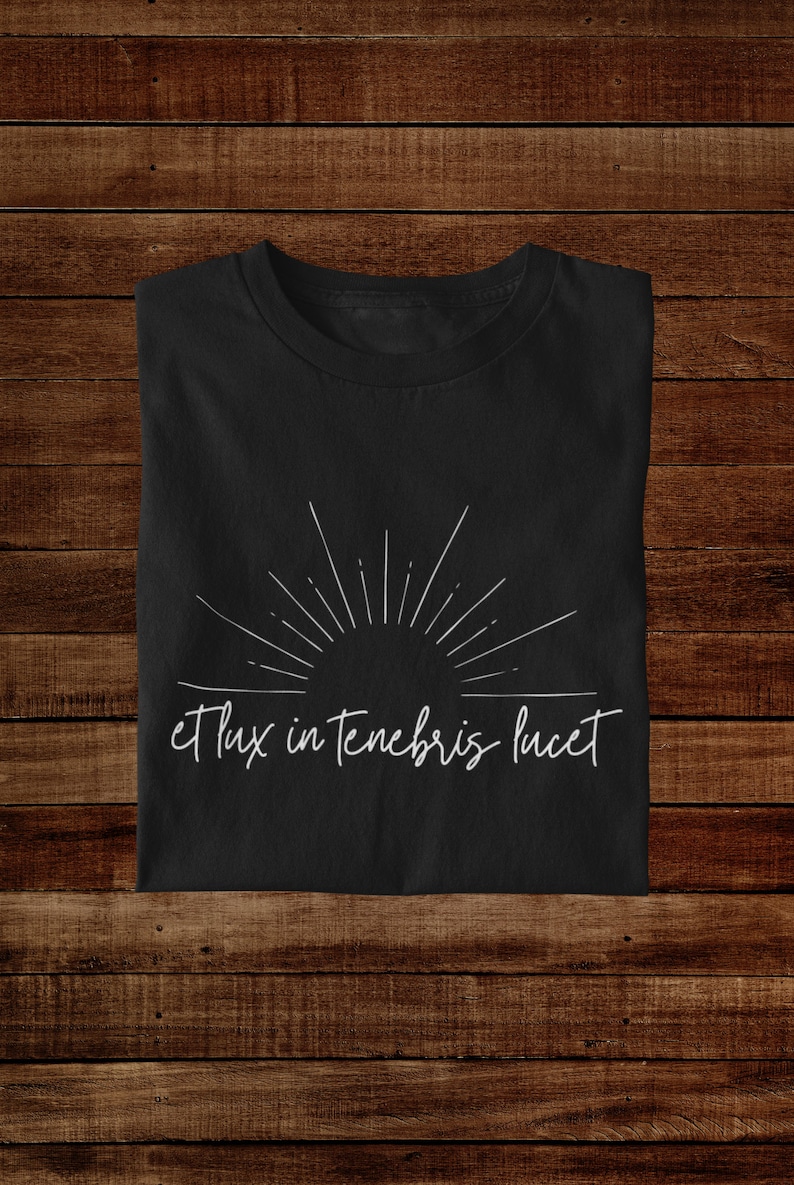 K&ouml;nnte beinhalten: Schwarzes T-Shirt mit einem wei&szlig;en Sonnenstrahlen-Design und dem Text "et lux in tenebris lucet" in handschriftlicher Schrift.