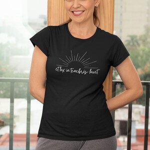 K&ouml;nnte beinhalten: Ein schwarzes T-Shirt mit einem wei&szlig;en Sonnenstrahl-Design und dem Text "et lux in tenebris lucet".