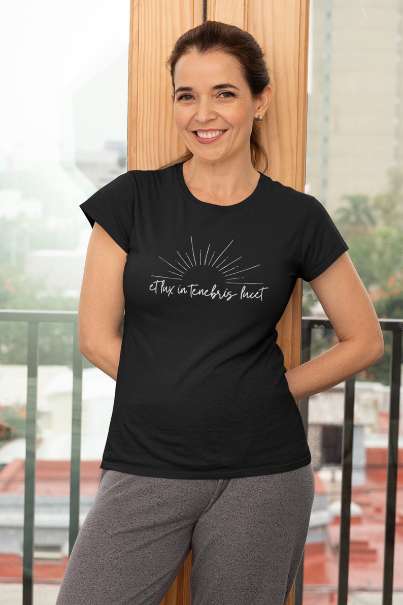 K&ouml;nnte beinhalten: Ein schwarzes T-Shirt mit einem wei&szlig;en Sonnenstrahl-Design und dem Text "et lux in tenebris lucet".