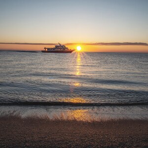 Puede incluir: Una vista panorámica de un barco navegando en el océano al atardecer. El sol, un orbe naranja brillante, proyecta un brillo dorado sobre el agua y la playa de arena en primer plano. El cielo cambia de azul a naranja.