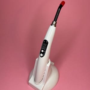 Könnte beinhalten: Weißes Dental-Härtungslicht mit roter Spitze und digitalem Display. Das Licht befindet sich in einem weißen Ständer.