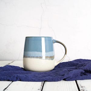 May include: A ceramic mug with a rounded shape and a handle. The mug features a gradient of colors, with a light blue top, a white middle section, and a speckled beige base. The handle has a metallic gold finish.