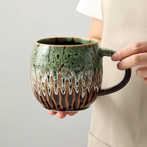May include: A round ceramic mug featuring a reactive glaze with a speckled mint green top that transitions into a dark brown base. The surface has a vertical ribbed texture with white drip-like patterns running down the middle section of the mug.