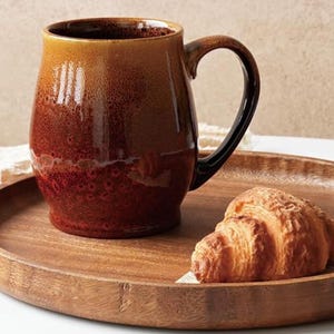 May include: A large, brown ceramic mug with a black handle sits on a wooden tray next to a croissant. The mug has a gradient color, transitioning from a lighter brown at the top to a darker, reddish-brown at the bottom. The tray is round.
