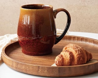 Può includere: Una grande tazza in ceramica marrone con manico nero è su un vassoio di legno con un croissant. La tazza ha una sfumatura di colore, che va dal marrone chiaro in alto al marrone rossastro scuro in basso. Il vassoio è rotondo.
