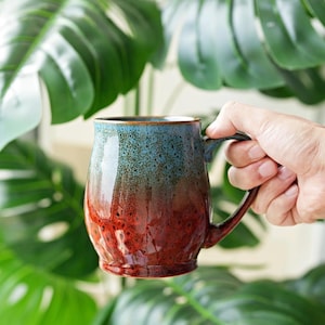 May include: A ceramic mug featuring a reactive glaze with a gradient transition from a speckled blue top to a deep red base. The mug has a rounded body, a curved handle, and a textured surface finish that displays variations in color and tone.