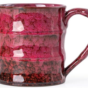 May include: A handmade ceramic mug with a unique, textured surface and a deep burgundy glaze. The mug features a large, curved handle and a capacity suitable for coffee or tea. The mug's design includes horizontal bands of color.
