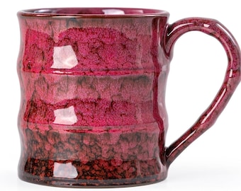 May include: A handmade ceramic mug with a unique, textured surface and a deep burgundy glaze. The mug features a large, curved handle and a capacity suitable for coffee or tea. The mug's design includes horizontal bands of color.