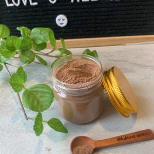 May include: A glass jar filled with a brown powder, with a gold lid and a wooden spoon with a scoop of the powder. The spoon has the text "@hibiscus_mantra" engraved on it.