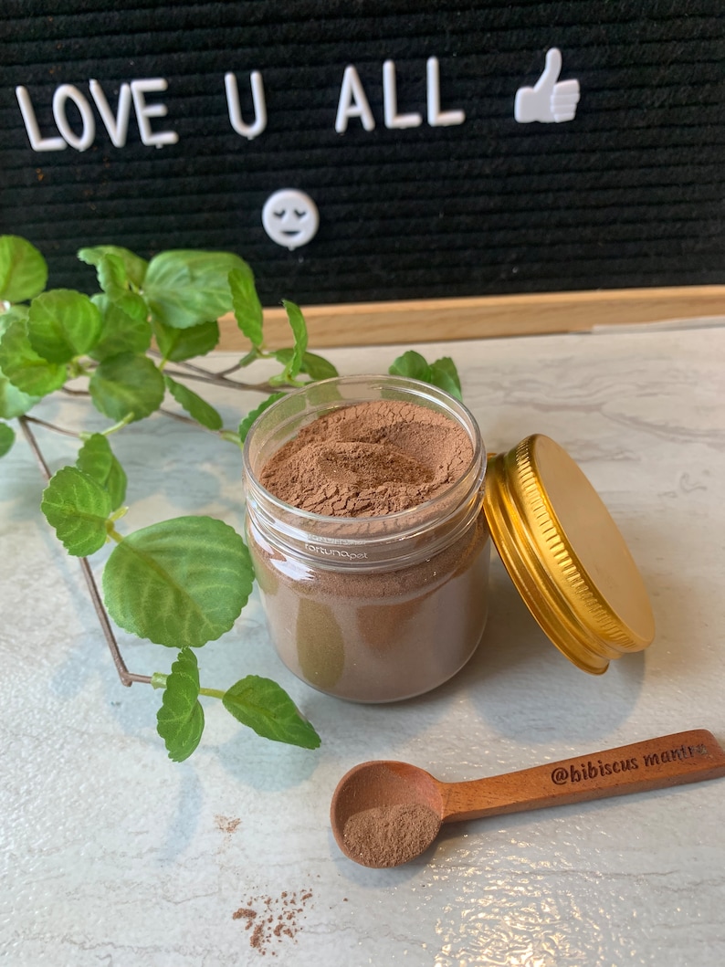 May include: A glass jar filled with a brown powder, with a gold lid and a wooden spoon with a scoop of the powder. The spoon has the text "@hibiscus_mantra" engraved on it.