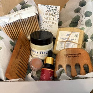 May include: A gift box filled with self-care items, including a wooden comb, a jar of body butter, a small jar of lip balm, a bottle of essential oil, a bar of soap, and a wooden scalp massager. The box is lined with white tissue paper and has a green and white floral pattern. The text on the soap reads "Carrot Orange Lemon Wild Turmeric De Body". The text on the scalp massager reads "Scalp massages are t-hair-apeutic".
