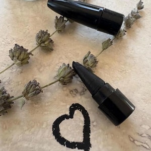 May include: A black cosmetic crayon with a removable cap, positioned next to a dried lavender sprig. A hand-drawn heart shape in black pigment is visible on the surface, demonstrating the color and texture of the cosmetic product.