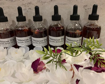 May include: Six clear glass bottles with black droppers filled with a reddish-brown liquid. Each bottle has a label that reads "Hibiscus Organic Ayurvedic Herbal Hair Growth". The bottles are surrounded by white flower petals and greenery.