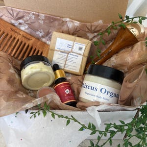 May include: A gift box filled with natural beauty products, including a wooden comb, a bar of soap, a jar of body butter, a bottle of oil, and a wooden gua sha tool. The box is lined with white tissue paper and decorated with green leaves. The products are labeled "Hibiscus Organic" and feature a hibiscus flower design.
