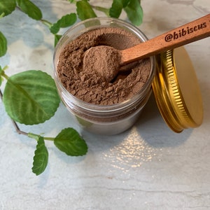 May include: A glass jar filled with a brown powder, with a wooden spoon resting on top of the powder. The spoon has the text "@bibiscus" engraved on it. The jar has a gold lid.