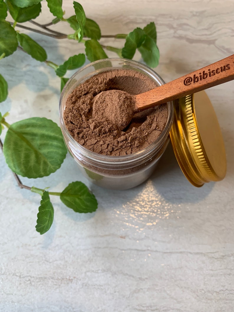 May include: A glass jar filled with a brown powder, with a wooden spoon resting on top of the powder. The spoon has the text "@bibiscus" engraved on it. The jar has a gold lid.