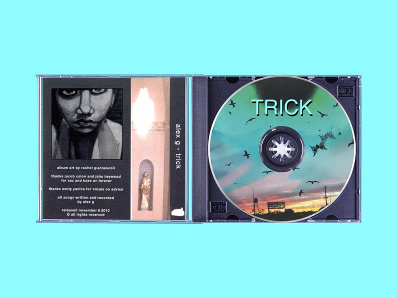 K&ouml;nnte beinhalten: Ein CD-Geh&auml;use mit einem Schwarzwei&szlig;bild einer Person auf dem vorderen Cover und einer blauen und gr&uuml;nen CD mit dem Text "TRICK" und einer Sonnenuntergangsszene auf dem hinteren Cover. Der Text "alex g - trick" ist auf dem R&uuml;cken des Geh&auml;uses aufgedruckt.