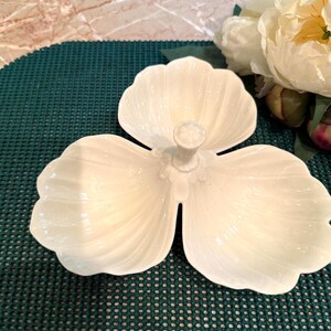 May include: A cream-colored ceramic serving dish shaped like a three-leaf clover or flower, featuring a central handle with a small knob. The dish has a glossy finish and scalloped edges, with embossed lines radiating from the center of each petal.