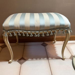 May include: An upholstered bench with a cream-colored frame and curved legs. The seat features vertical stripes in light blue and cream, trimmed with a decorative fringe and tassels. A classic piece of furniture.