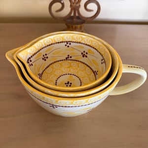 May include: Nested yellow ceramic measuring cups with a handle. The cups have a decorative floral pattern in yellow and brown. The cups are stacked inside each other. The background is a light brown surface.