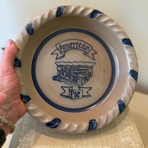 May include: A tan ceramic pie plate with a blue rim and accents. The center features a blue illustration of a pie with the words "American Pie" in a banner. The plate has a decorative, twisted edge.