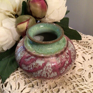 May include: A small, hand-crafted ceramic vase with a green glaze and pink and white accents. The vase is sitting on a white crocheted doily.