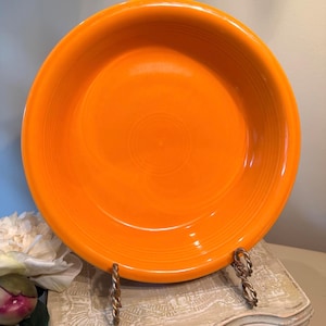 May include: A bright orange ceramic bowl with a smooth, glossy finish and a wide, circular rim. The bowl features a series of concentric, raised ring patterns etched into the interior surface, creating a subtle texture around the center.