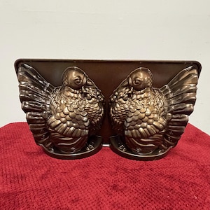 May include: A bronze-colored metal turkey-shaped baking pan. The pan features two detailed turkey molds with textured feathers and bodies. The pan is designed for baking and has a rectangular shape.